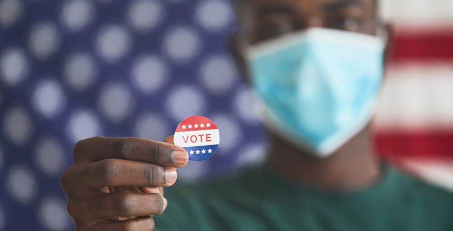 A man wearing a surgial mask standing in front of an American flag holds up a 'vote' sticker.