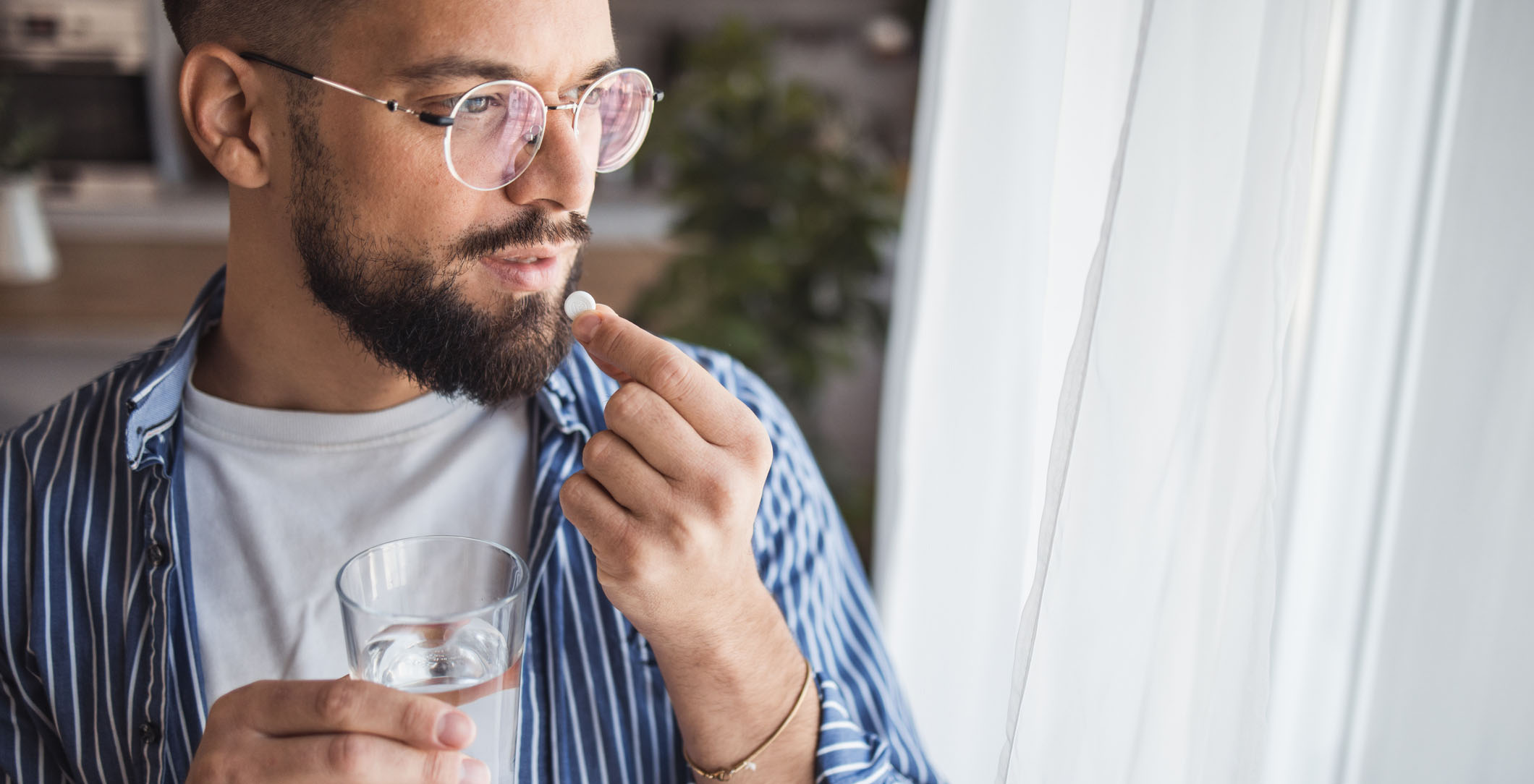 Young man standing by a window, with a glass of water in one hand, holding a pill close to his mouth with the other hand.